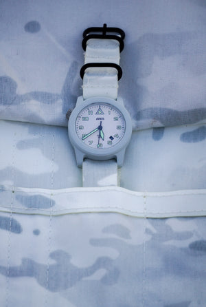 Close-up of a Cerakote Snow White Field-1 timer watch featuring a lumed ARES arrow crown on a Multicam Alpine white camouflage-patterned strap in a matching Alpine 4-slot ARES watch roll 